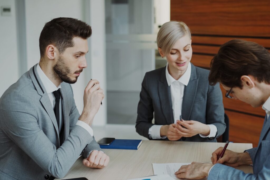 Young businessman in suit signing contract with his colleague while corporate lawyer explaining details of partnership sitting in modern office indoors